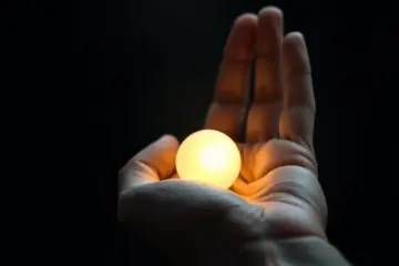 A weathered hand gently cradles a small, glowing orb of warm light against a dark, blurred background.