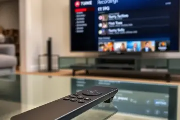 A sleek remote on a glass table, with a TV in the background displaying an electronic program guide for scheduled recordings.